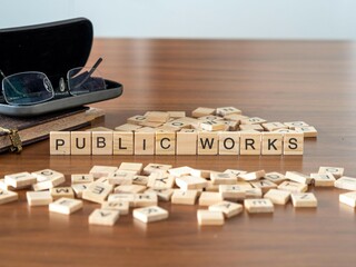 public works word or concept represented by wooden letter tiles on a wooden table with glasses and a book - obrazy, fototapety, plakaty