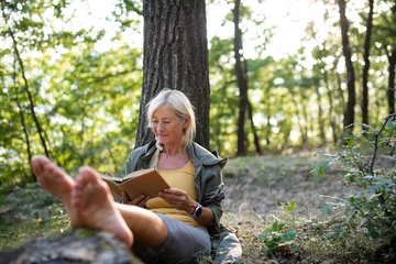 Rolgordijnen Senior woman relaxing and reading book outdoors in forest. © Halfpoint