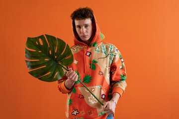 Obraz premium Fashion studio portrait of a young man in hoodie with big leaf posing over orange background.