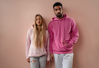 Fashion studio portrait of young biracial couple in hoodies posing over pink background.