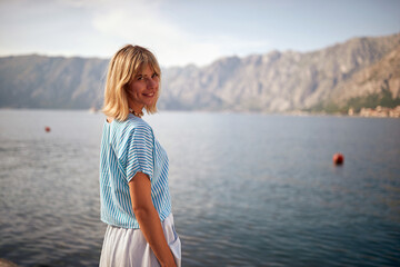Cute blonde woman smiling, standing on pier in front of beautiful view of water, mountains in background. Travel, lifestyle, holiday, summertime concept.