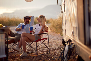Couple at sunset cheering with drinks. Sitting in front of camper rv. Fun, togetherness, travel,...