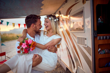Newlyweds in boho style in front of decorated camper rv. Groom holding bride in his arms. Wedding...