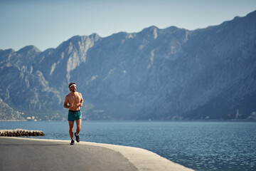Sporty man on a run by water on jetty. Beautiful background of sea and mountains. Morning run. Sport, vitality, recreation concept.