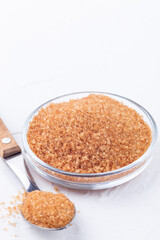 Brown sugar crystals or cane sugar in glass bowl and in spoon on white background, vertical, copy space