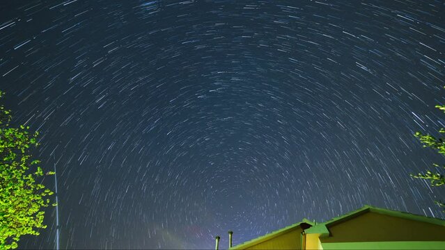 The stars move around the North Star. Trees and the roof of a house in the foreground. Time lapse of star trails in the night sky in Italy.