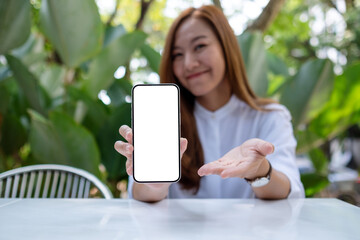 Mockup image of a young woman holding and showing a mobile phone with blank white screen in the...