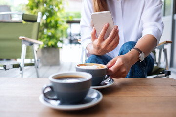 Closeup image of a young woman holding and using mobile phone while drinking coffee with friend in cafe