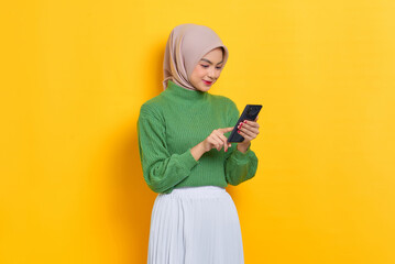 Smiling beautiful Asian woman in green sweater using mobile phone received good news isolated over yellow background