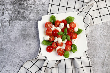 Italian caprese salad with sliced tomatoes, mozzarella, basil, olive oil on a white square  plate on a dark background. Top view, flat lay