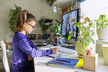 Girl child sitting at home at table with computer listening online lesson