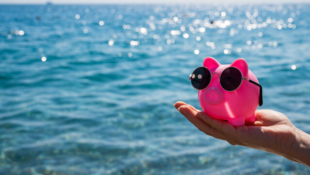 A woman holds a piggy bank in sunglasses on the background of the sea. Budget vacation. - Powered by Adobe
