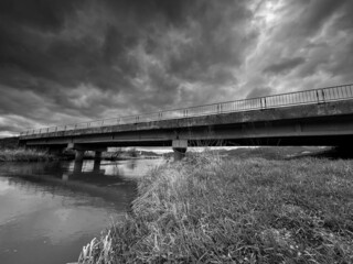 Wide angle monochrome image, concrete bridge over Olt river, forming dark clouds on the sky.