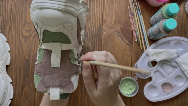Female's hands dip brush in green paint and apply it to sneaker. Palette, tools and tubes on wooden table. Hobbies, manual work and leisure at home. Design of sports shoes using acrylic.