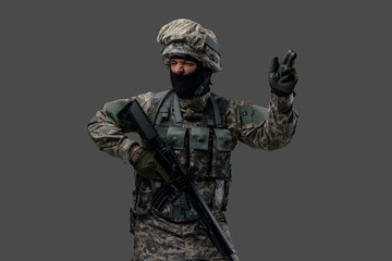 Proud serviceman dressed in camouflage clothes with rifle gesturing against grey background.