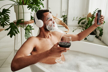 Young man with facial mask drinking wine and making selfie portrait on mobile phone while taking a...