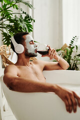 Young man in mask drinking red wine and listening to music in headphones while relaxing in bath
