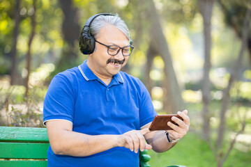 Happy Indian or asian senior man wearing headphones watching videos on his smart phone while sitting on the bench at park , Old grey hair male wearing glasses using android mobile phone outdoor.