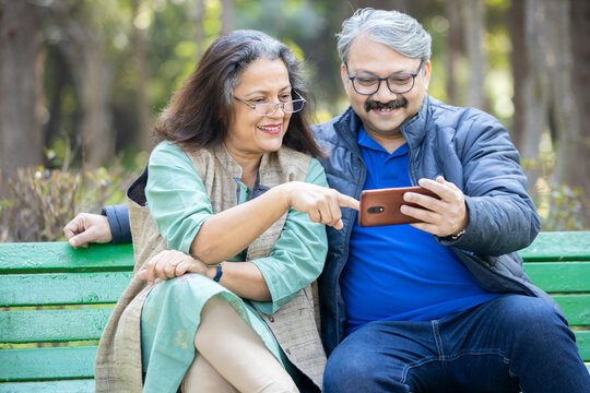 Happy Indian Asian Senior Couple Using Smart Phone At Park, Old Man And Woman Watching Video Social Media On Mobile Phone While Sitting On Bench In The Garden.