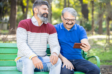 Two happy indian senior men watching video or social media on smart android phone while sitting at...