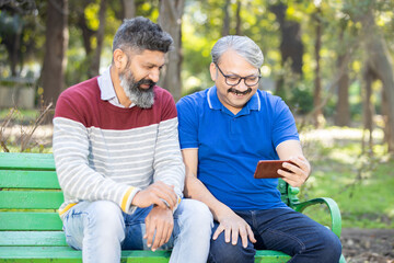 Fototapeta premium Two happy indian senior men watching video or social media on smart android phone while sitting at park outdoor, Mature old people using technology, Friends having fun.