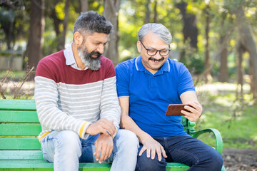 Two happy indian senior men watching video or social media on smart android phone while sitting at park outdoor, Mature old people using technology, Friends having fun.