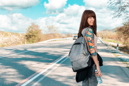 A young pretty caucasian woman walks along the countryside road with a backpack in her hands. Back view. Copy space. Concept of hitchhiking and local travel