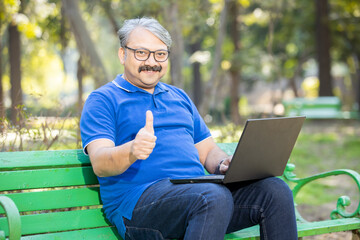 Portrait of happy Indian asian senior man sitting on bench using laptop at park looking at camera...