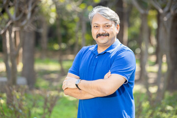 Happy senior indian asian man with arms crossed standing at park outdoor, Old mustache and grey haired male smiling wearing blue t-shirt. Copy space. - Powered by Adobe