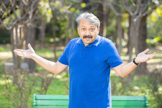 Portrait of Indian Asian Senior man with arms and hands raised at park outdoor. clueless and confused expression.