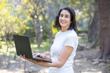 Portrait of mature Indian asian woman using laptop in the park outdoors, social media portable computer working, technology concept. looking at camera