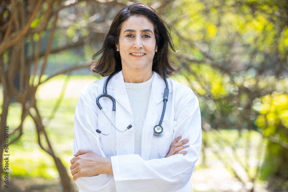 Wall mural Portrait smiling female indian doctor wearing apron and stethoscope with cross arm standing outdoor in the park, healthcare profession.