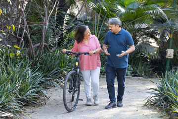 Happy Indian asian senior couple walking their bike talking in park.mature couple in summer park.Retired man and old woman with bicycles outside nature. - Powered by Adobe