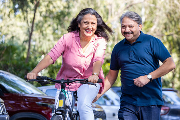 Fototapeta na wymiar Happy Indian Senior couple riding bicycle in the park summer, active old age people and lifestyle. Elderly woman learn to ride cycle with man. retired people having enjoy life. selective focus.