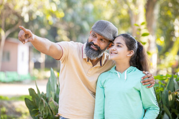 Happy indian mature father pointing finger and sowing something to his daughter at distance in the park. single parent.