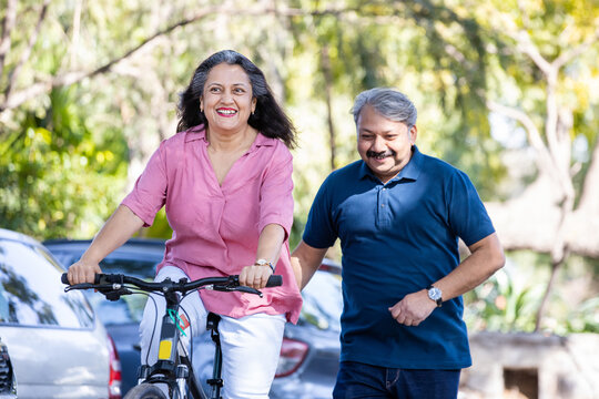 Happy Indian Senior Couple Riding Bicycle In The Park Summer, Active Old Age People And Lifestyle. Elderly Woman Learn To Ride Cycle With Man. Retired People Having Enjoy Life. Selective Focus.