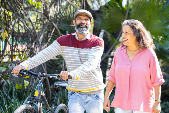 Happy Indian Asian Senior Couple Walking Their Bike Talking In Park.mature Couple In Summer Park.Retired Man And Old Woman With Bicycles Outside Nature.