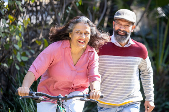 Happy Indian Senior Couple Riding Bicycle In The Park Summer, Active Old Age People And Lifestyle. Elderly Woman Learn To Ride Cycle With Man. Retired People Having Enjoy Life. Selective Focus.