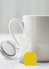 Teabag with yellow blank tag of black tea next to porcelain cup on white with infuser strainer.