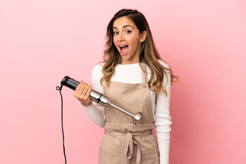 Young woman using hand blender over isolated pink background with surprise facial expression