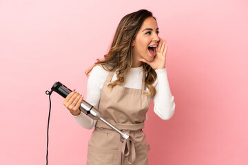 Young woman using hand blender over isolated pink background shouting with mouth wide open to the side