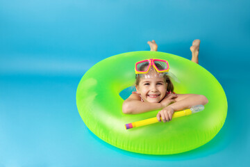 Happy little child girl in swimming mask and snorkel lying on a bright inflatable circle and smiling. Blue background. Summer and vacation concept