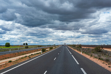 Truck driving along a straight highway with a flat horizon and a cloudy sky in the community of La Mancha, Spain.