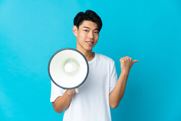 Young Chinese man isolated on blue background shouting through a megaphone and pointing side