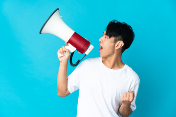 Young Chinese man isolated on blue background shouting through a megaphone to announce something in...