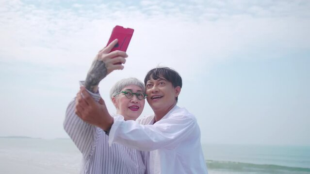 Asian retired couple relaxing by the sea in summer. A senior woman taking a selfie photo with her partner on the beach.