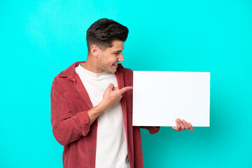 Young handsome caucasian man isolated on blue bakcground holding an empty placard with happy expression and pointing it