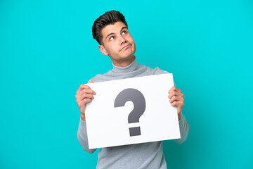 Young handsome caucasian man isolated on blue bakcground holding a placard with question mark symbol and thinking