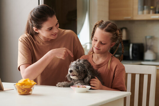 Smiling mother and daughter letting dog to eat from bowl on kitchen table