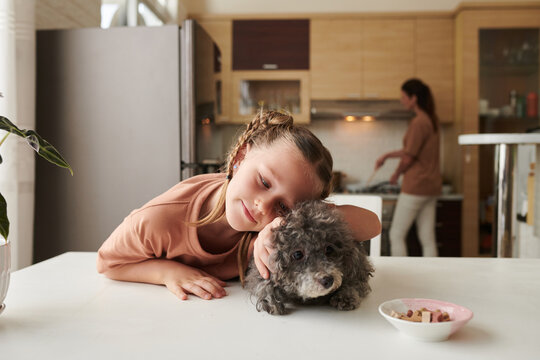 Smiling girl patting and hugging small dog trying to steal treat from bowl on kitchen table